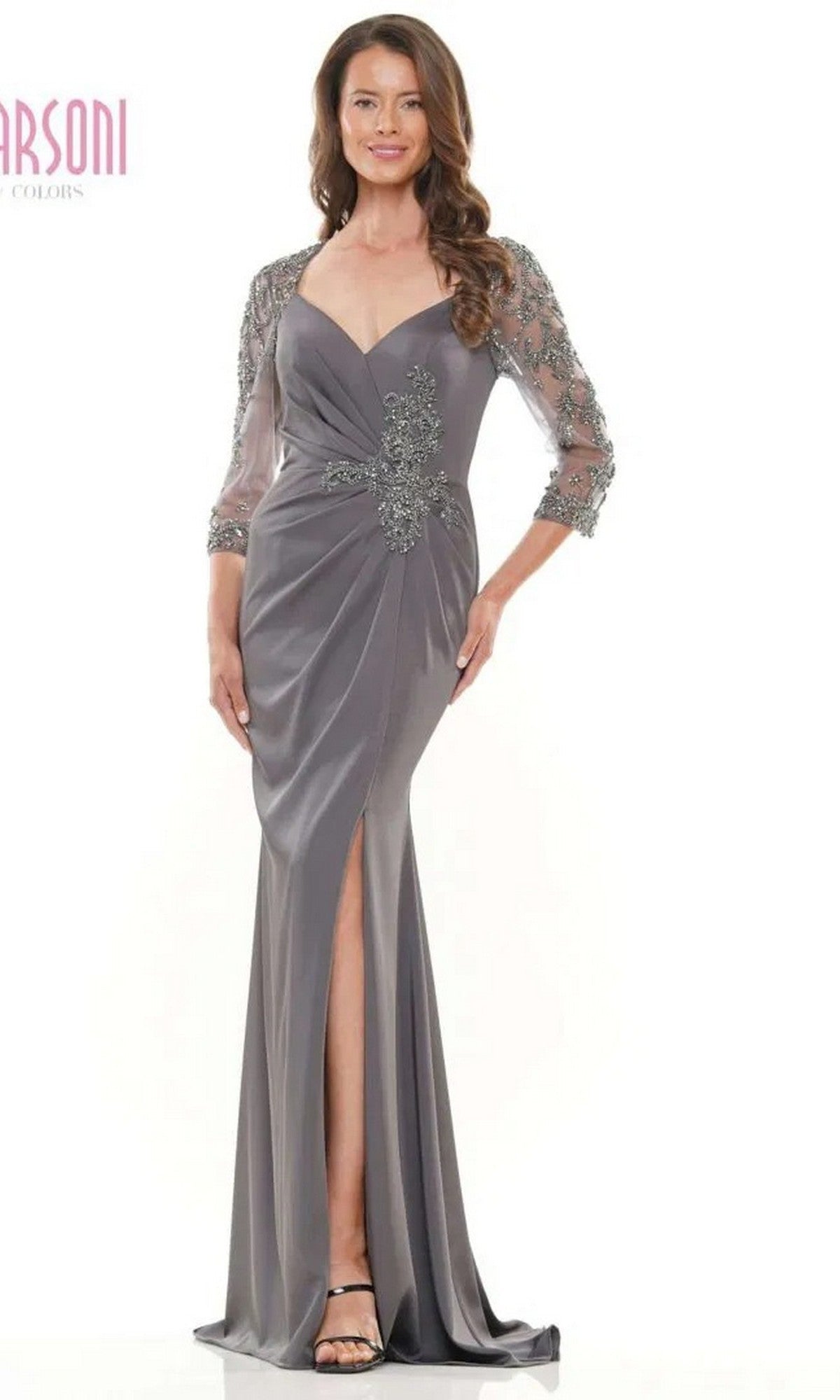 A woman wearing the Marsoni MV1070 Long Formal Dress in charcoal with beaded appliques, sheer sleeves, and a front slit stands smiling.